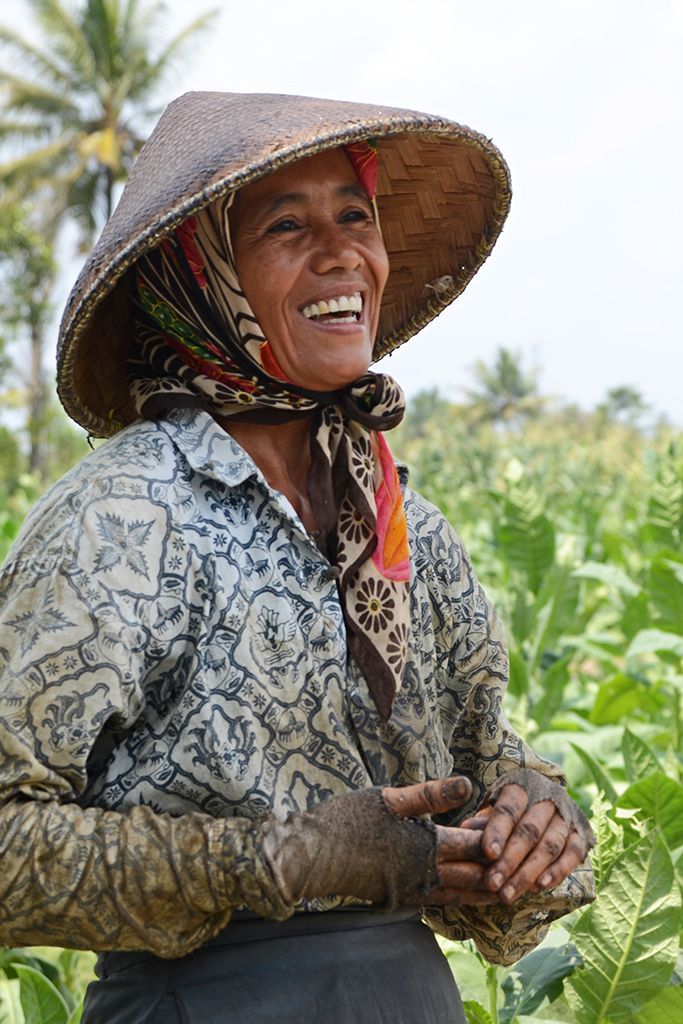 An Indonesian farmer in a woven hat and batik shirt, smiling in a field